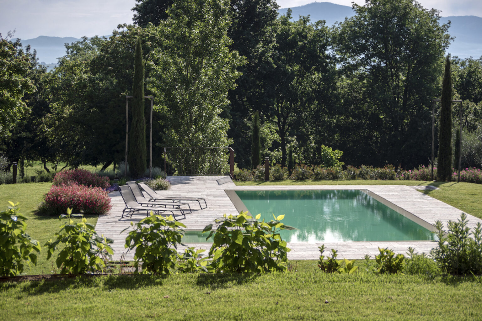 Serene outdoor pool surrounded by lush greenery and landscaped gardens, featuring comfortable lounge chairs and vibrant flowers, ideal for relaxation and leisure. | Only on SopranoVillas