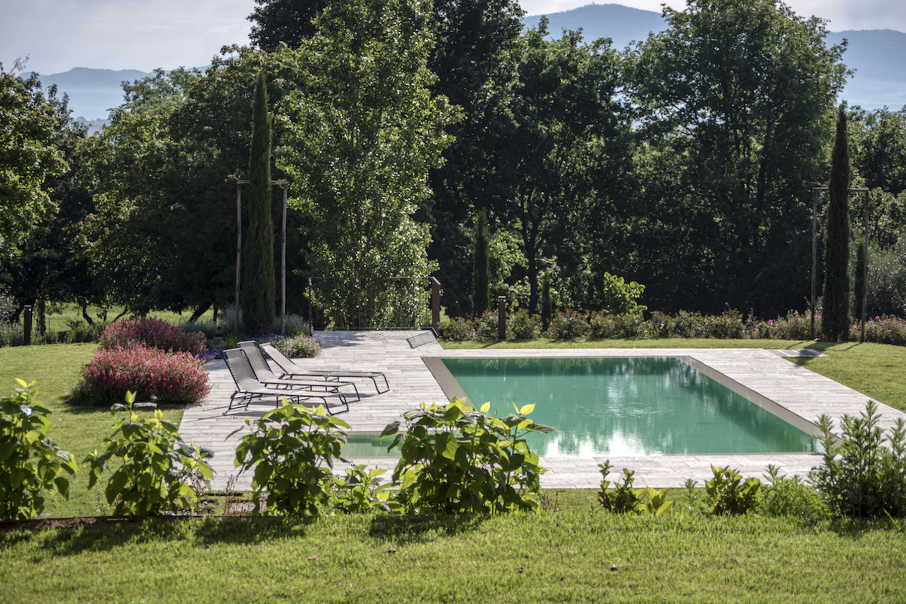 Serene outdoor pool surrounded by lush greenery and landscaped gardens, featuring comfortable lounge chairs and vibrant flowers, ideal for relaxation and leisure. | Only on SopranoVillas