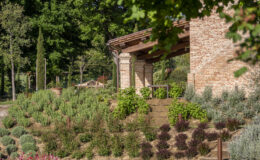Rustic brick building surrounded by lush greenery and landscaped gardens, featuring a variety of plants, shrubs, and trees in a serene outdoor setting.