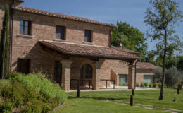 Exterior view of a rustic brick house with a tiled roof, featuring large windows and a covered porch area. Surrounded by well-maintained gardens and greenery, the property showcases a blend of traditional architecture and natural beauty.