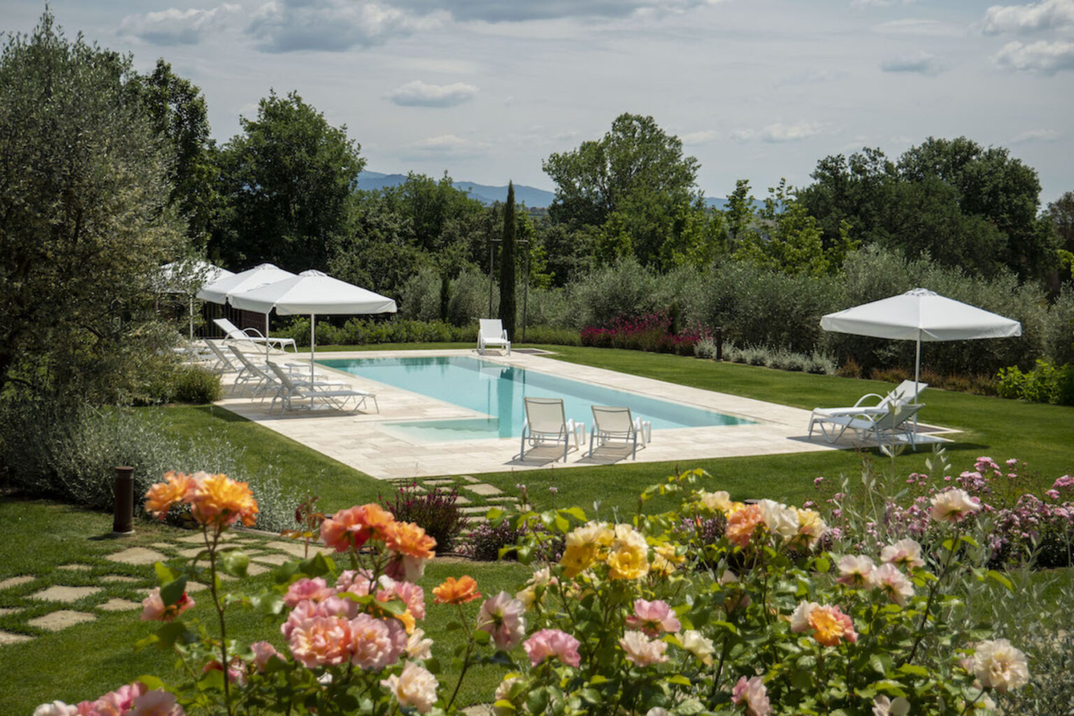 Beautiful outdoor swimming pool surrounded by lush greenery and blooming flowers, featuring white lounge chairs and umbrellas for relaxation on a sunny day. Perfect for a tranquil getaway or a luxurious retreat. | Only on SopranoVillas