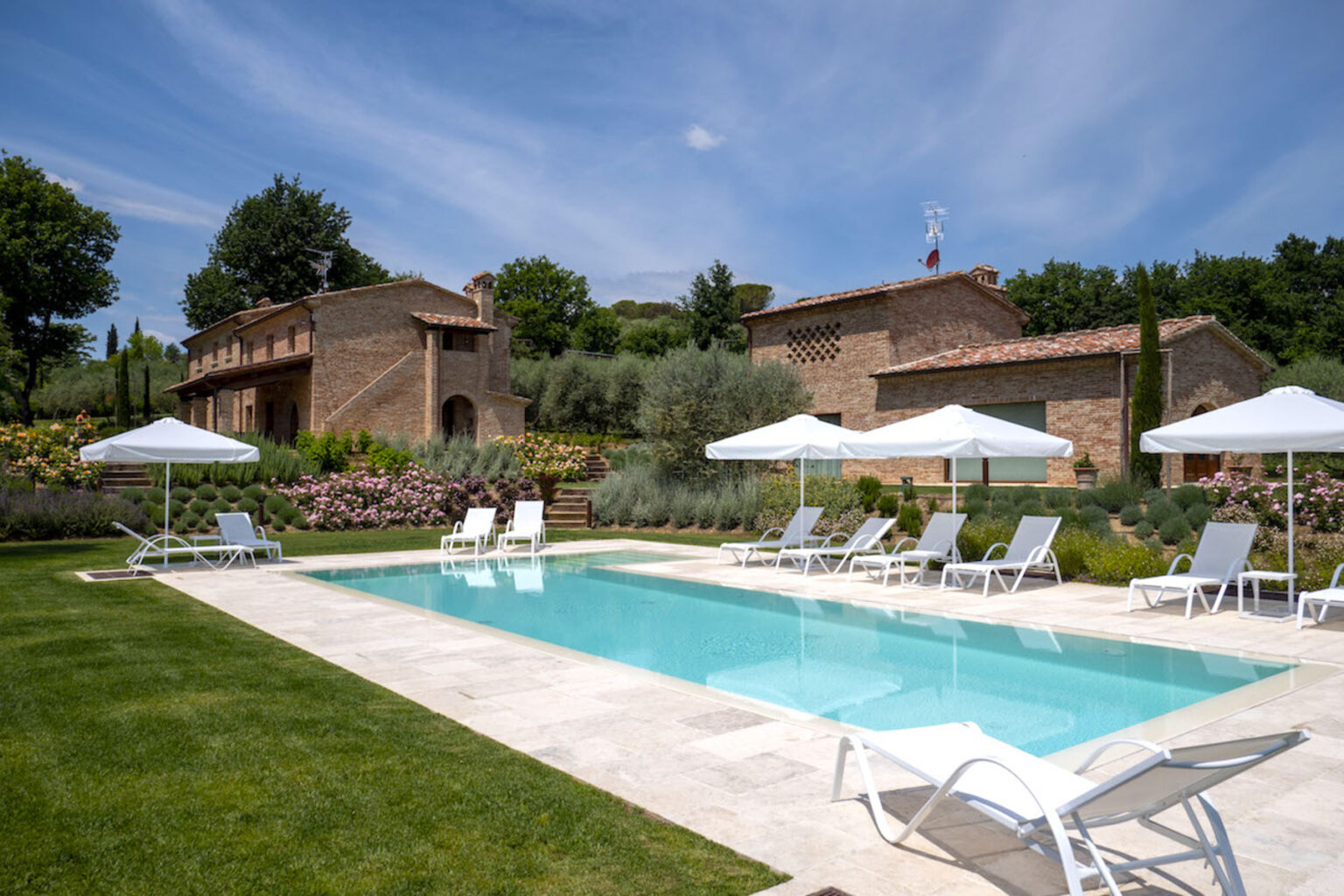 Luxurious outdoor swimming pool surrounded by sun loungers and umbrellas, with a picturesque stone villa and lush gardens in the background, under a clear blue sky. | Only on SopranoVillas
