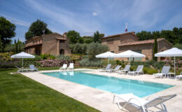 Luxurious outdoor swimming pool surrounded by sun loungers and umbrellas, with a picturesque stone villa and lush gardens in the background, under a clear blue sky.