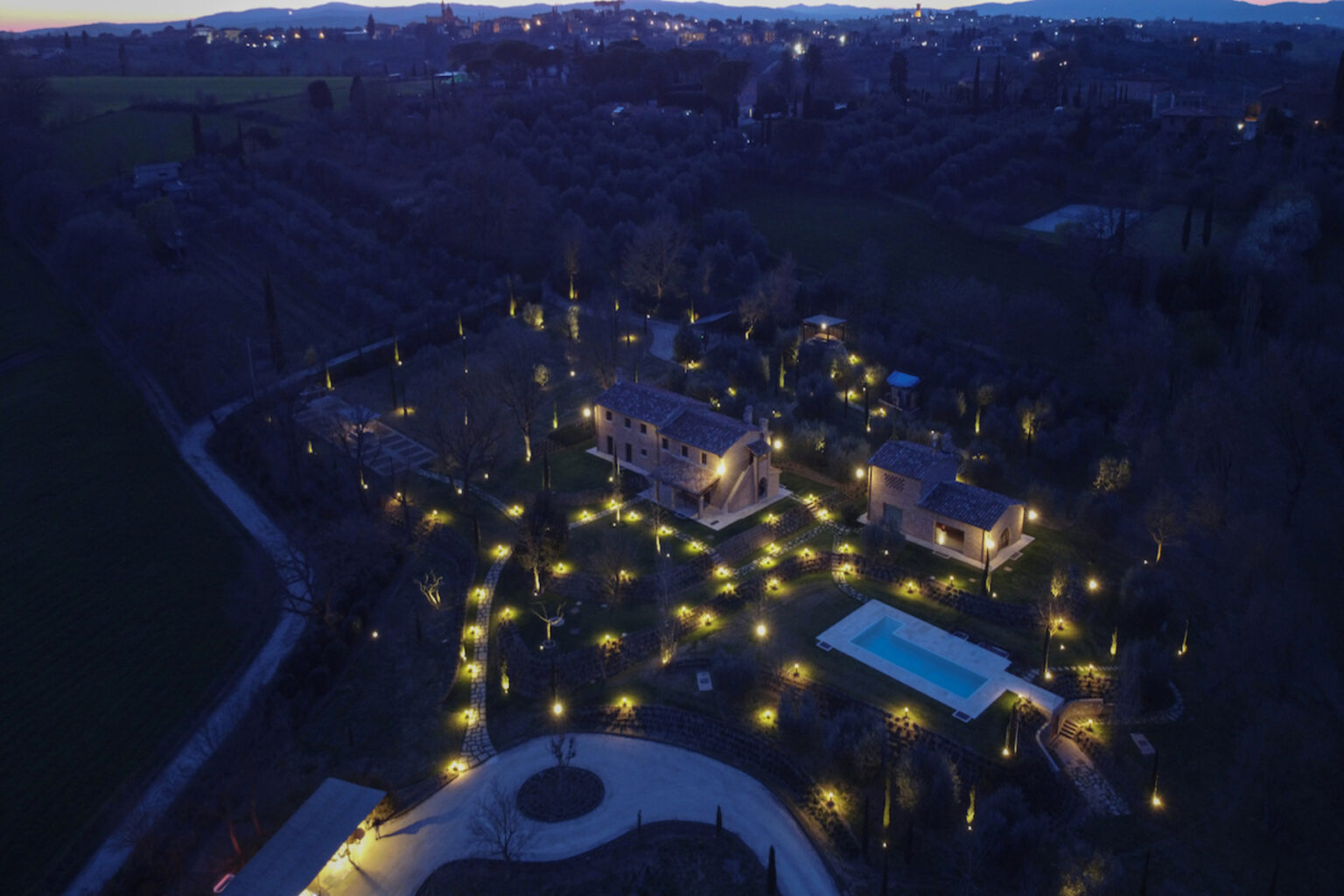 Aerial view of a beautifully illuminated property at night, featuring two houses, a swimming pool, and a landscaped garden adorned with decorative lights, set against a dark sky. | Only on SopranoVillas