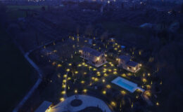 Aerial view of a beautifully illuminated property at night, featuring two houses, a swimming pool, and a landscaped garden adorned with decorative lights, set against a dark sky.