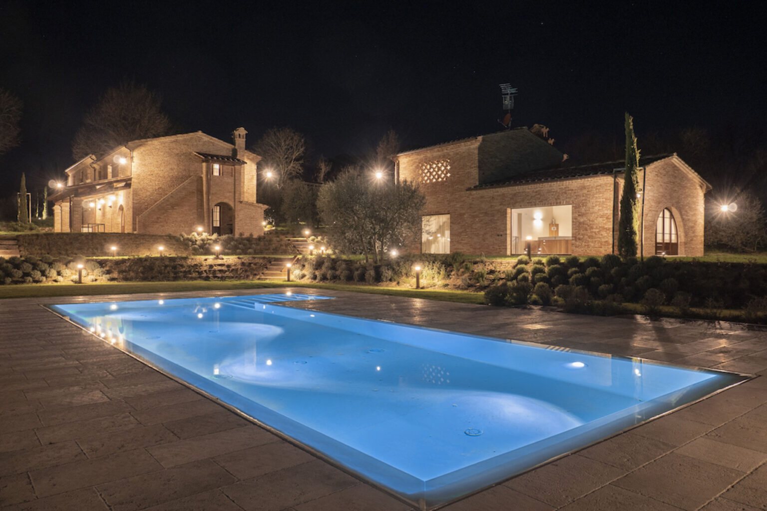Elegant night view of a luxurious villa featuring a glowing blue swimming pool, surrounded by landscaped gardens and softly lit pathways, showcasing modern architecture and serene outdoor ambiance. | Only on SopranoVillas