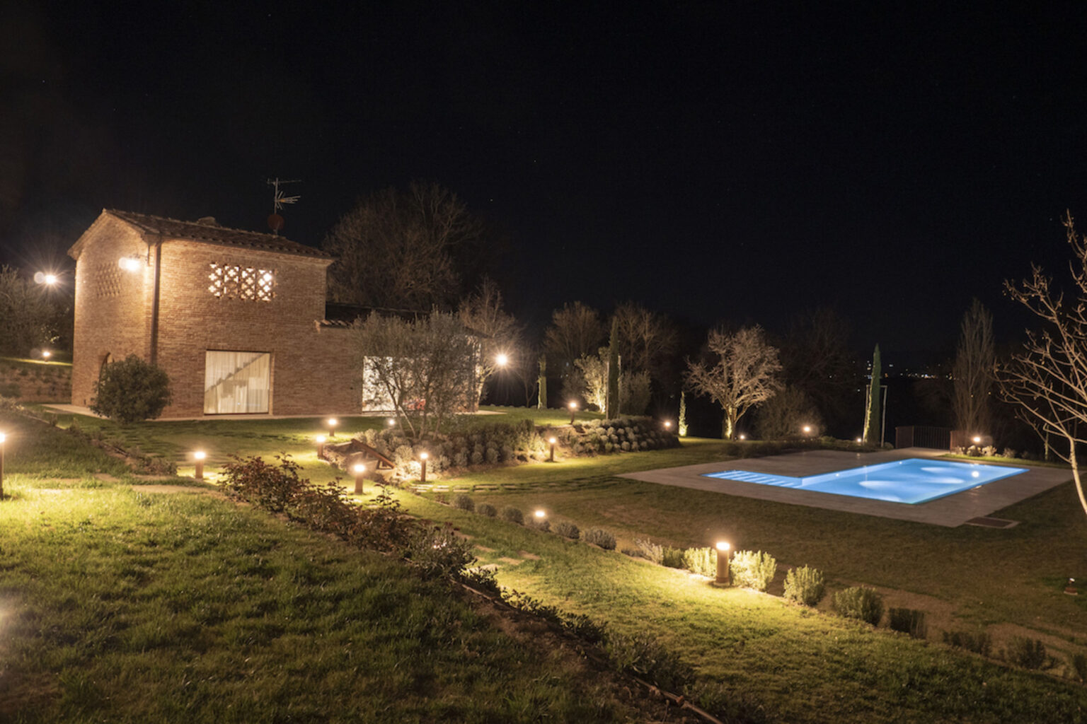 A beautifully illuminated countryside villa at night, featuring a brick exterior, landscaped gardens, and a serene swimming pool surrounded by soft lighting. | Only on SopranoVillas