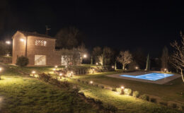 A beautifully illuminated countryside villa at night, featuring a brick exterior, landscaped gardens, and a serene swimming pool surrounded by soft lighting.