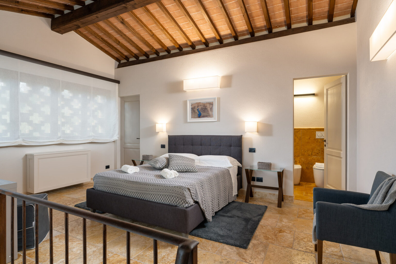 Modern bedroom interior featuring a comfortable bed, stylish decor, and natural light from large windows. The room includes a seating area and an en-suite bathroom, showcasing a blend of contemporary and rustic design elements. Ideal for a relaxing stay. | Only on SopranoVillas