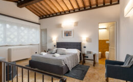 Modern bedroom interior featuring a comfortable bed, stylish decor, and natural light from large windows. The room includes a seating area and an en-suite bathroom, showcasing a blend of contemporary and rustic design elements. Ideal for a relaxing stay.
