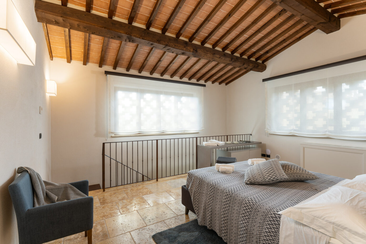 Cozy bedroom featuring a modern design with wooden beams, large windows, and stylish furnishings. The room includes a comfortable bed with patterned bedding, a gray armchair, and natural stone flooring, creating a warm and inviting atmosphere. Ideal for relaxation and comfort. | Only on SopranoVillas