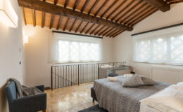 Cozy bedroom featuring a modern design with wooden beams, large windows, and stylish furnishings. The room includes a comfortable bed with patterned bedding, a gray armchair, and natural stone flooring, creating a warm and inviting atmosphere. Ideal for relaxation and comfort.