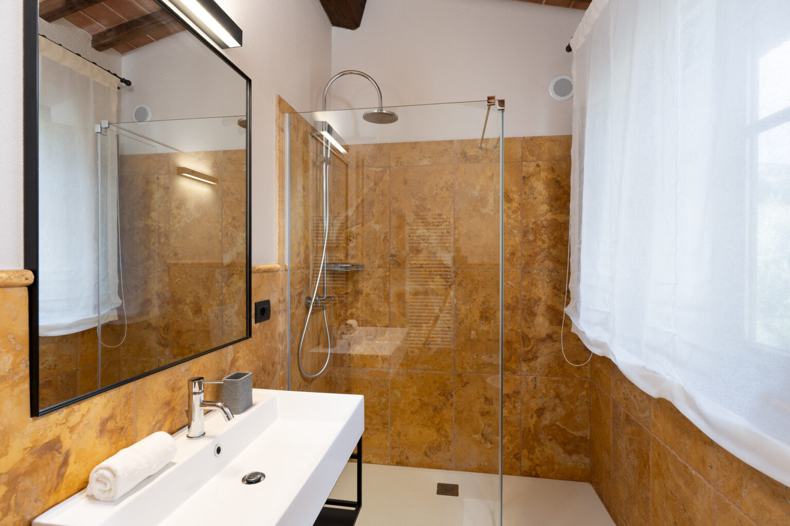 Modern bathroom featuring a walk-in shower with glass doors, a sleek white vanity with a mirror, and warm-toned marble walls. Natural light filters through sheer curtains, enhancing the contemporary design. | Only on SopranoVillas
