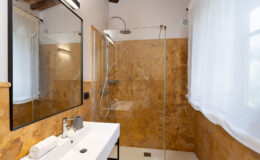 Modern bathroom featuring a walk-in shower with glass doors, a sleek white vanity with a mirror, and warm-toned marble walls. Natural light filters through sheer curtains, enhancing the contemporary design.