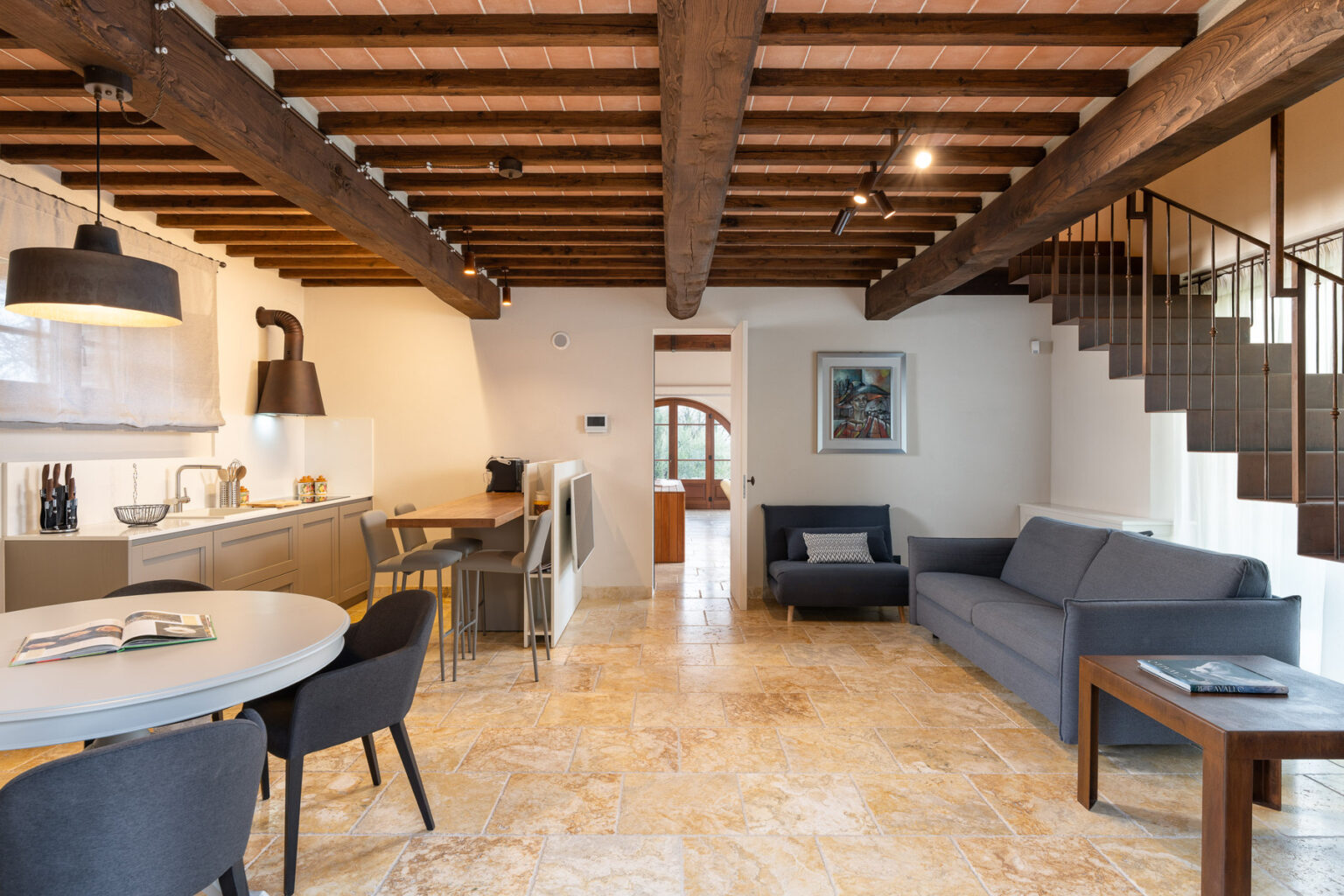 Modern living space featuring a stylish kitchen with wooden beams, a dining area with round table and chairs, and a cozy seating area with a gray sofa. The interior showcases natural stone flooring and warm lighting, creating a welcoming atmosphere. | Only on SopranoVillas
