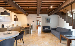 Modern living space featuring a stylish kitchen with wooden beams, a dining area with round table and chairs, and a cozy seating area with a gray sofa. The interior showcases natural stone flooring and warm lighting, creating a welcoming atmosphere.