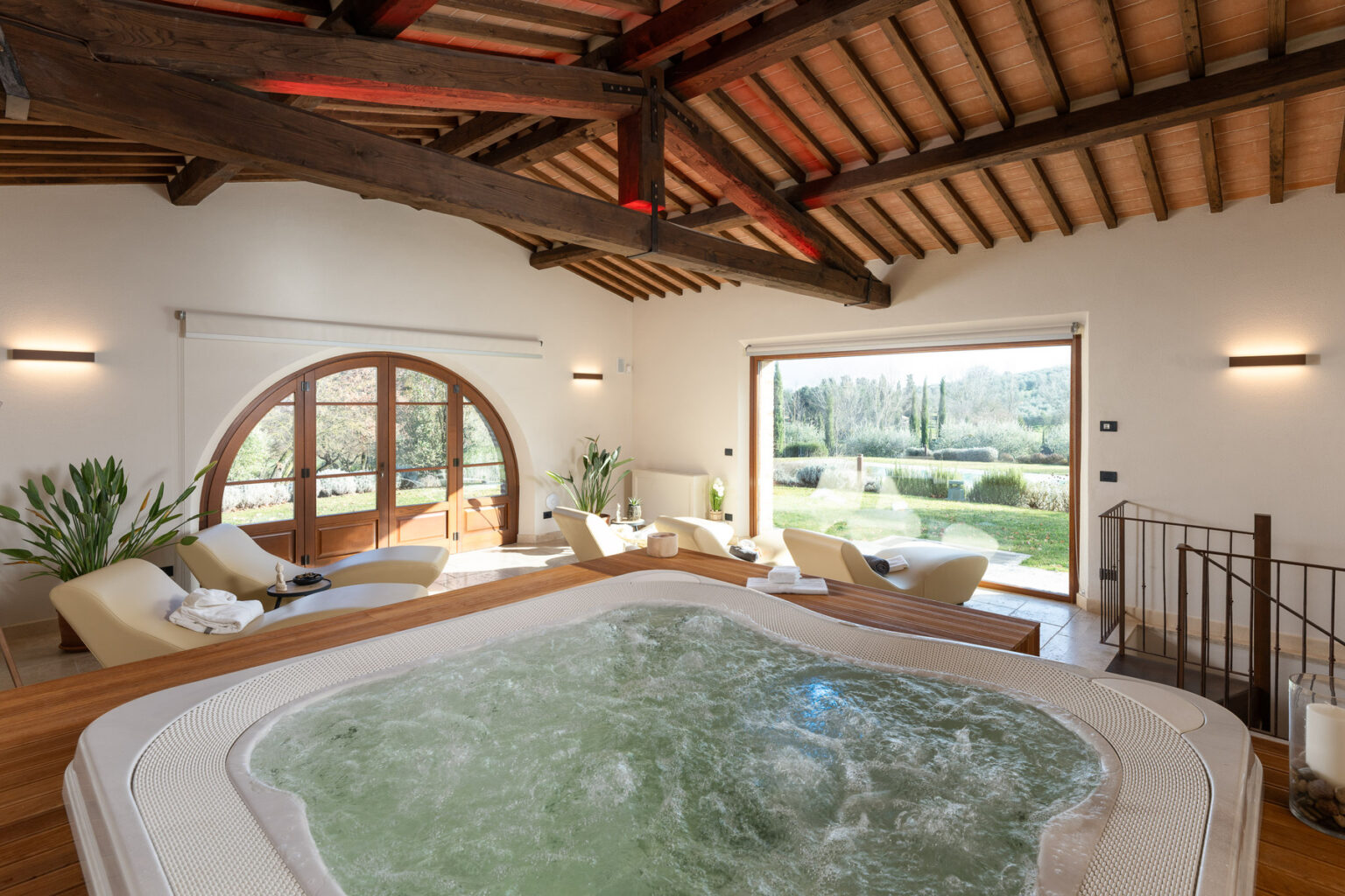 Luxurious indoor spa area featuring a jacuzzi, comfortable loungers, and large windows overlooking a scenic garden, with rustic wooden beams and warm lighting creating a relaxing atmosphere. | Only on SopranoVillas