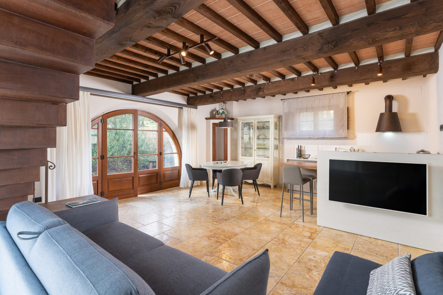 Spacious modern living area featuring wooden beams, a comfortable sofa, and a dining table with chairs, overlooking a large window with elegant doors. The open layout connects the living space to a stylish kitchen, showcasing a blend of contemporary and rustic design elements. | Only on SopranoVillas