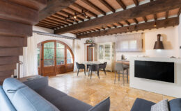 Spacious modern living area featuring wooden beams, a comfortable sofa, and a dining table with chairs, overlooking a large window with elegant doors. The open layout connects the living space to a stylish kitchen, showcasing a blend of contemporary and rustic design elements.