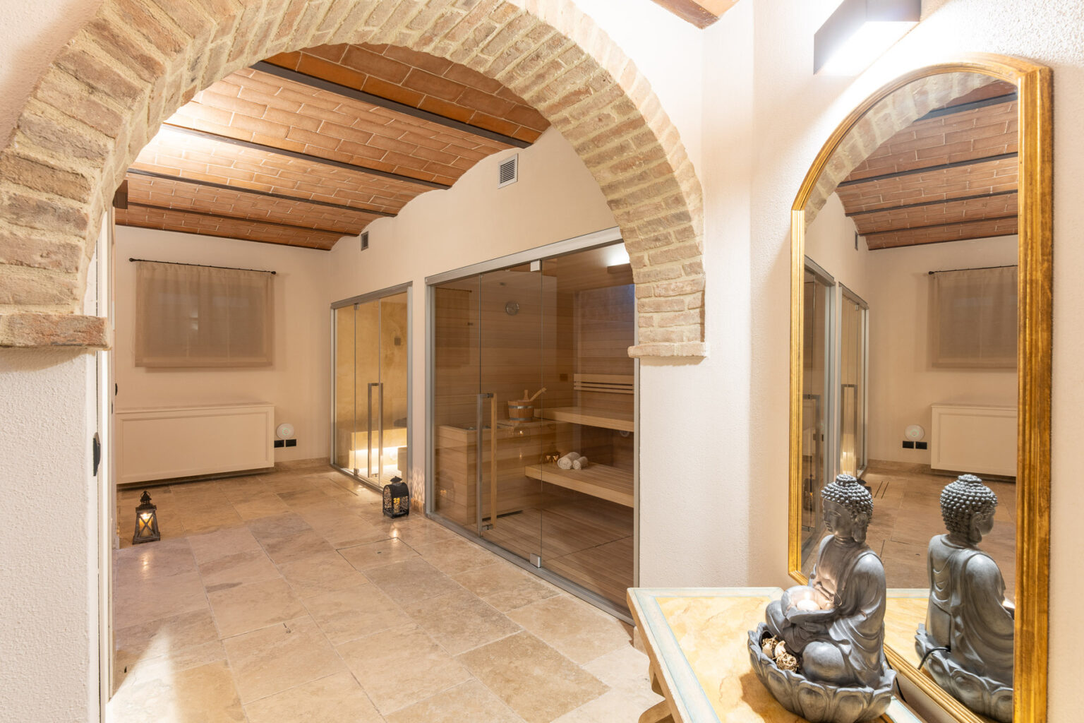 Serene spa interior featuring arched ceilings, stone flooring, and a reflection in a large mirror, with a sauna area visible in the background and decorative Buddha statues on a table. | Only on SopranoVillas