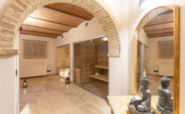 Serene spa interior featuring arched ceilings, stone flooring, and a reflection in a large mirror, with a sauna area visible in the background and decorative Buddha statues on a table.