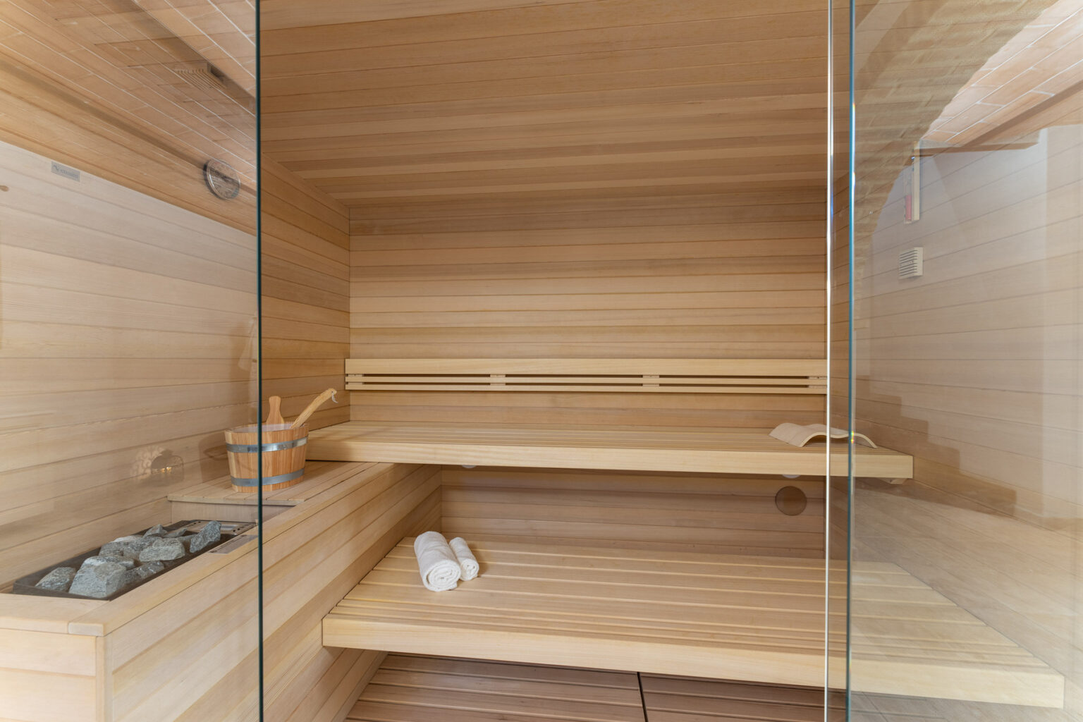 Interior of a modern wooden sauna featuring a glass door, wooden benches, and a stone heater, ideal for relaxation and wellness. | Only on SopranoVillas