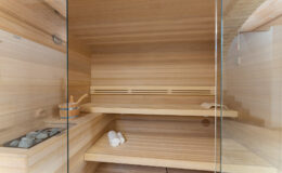 Interior of a modern wooden sauna featuring a glass door, wooden benches, and a stone heater, ideal for relaxation and wellness.