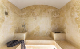 Luxurious bathroom interior featuring warm beige stone walls, a spacious shower area, and a modern faucet. Soft lighting enhances the elegant design, creating a serene atmosphere.