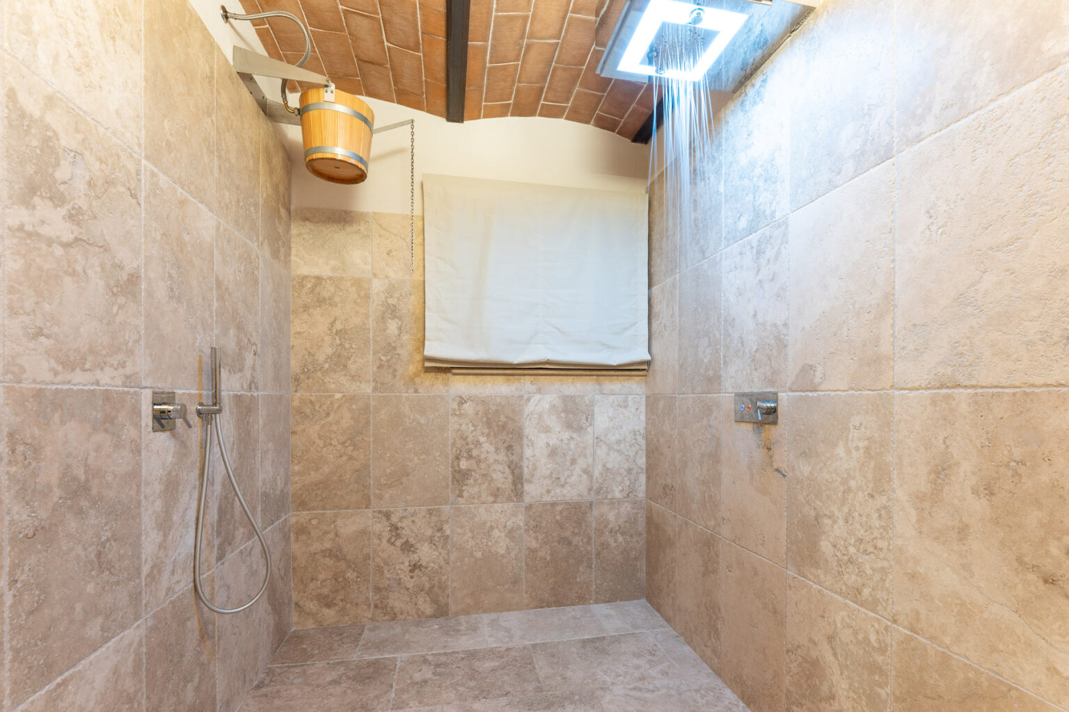 Spacious modern shower with beige stone tiles, featuring dual wall-mounted showerheads and natural light from a skylight. | Only on SopranoVillas