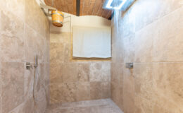 Spacious modern shower with beige stone tiles, featuring dual wall-mounted showerheads and natural light from a skylight.