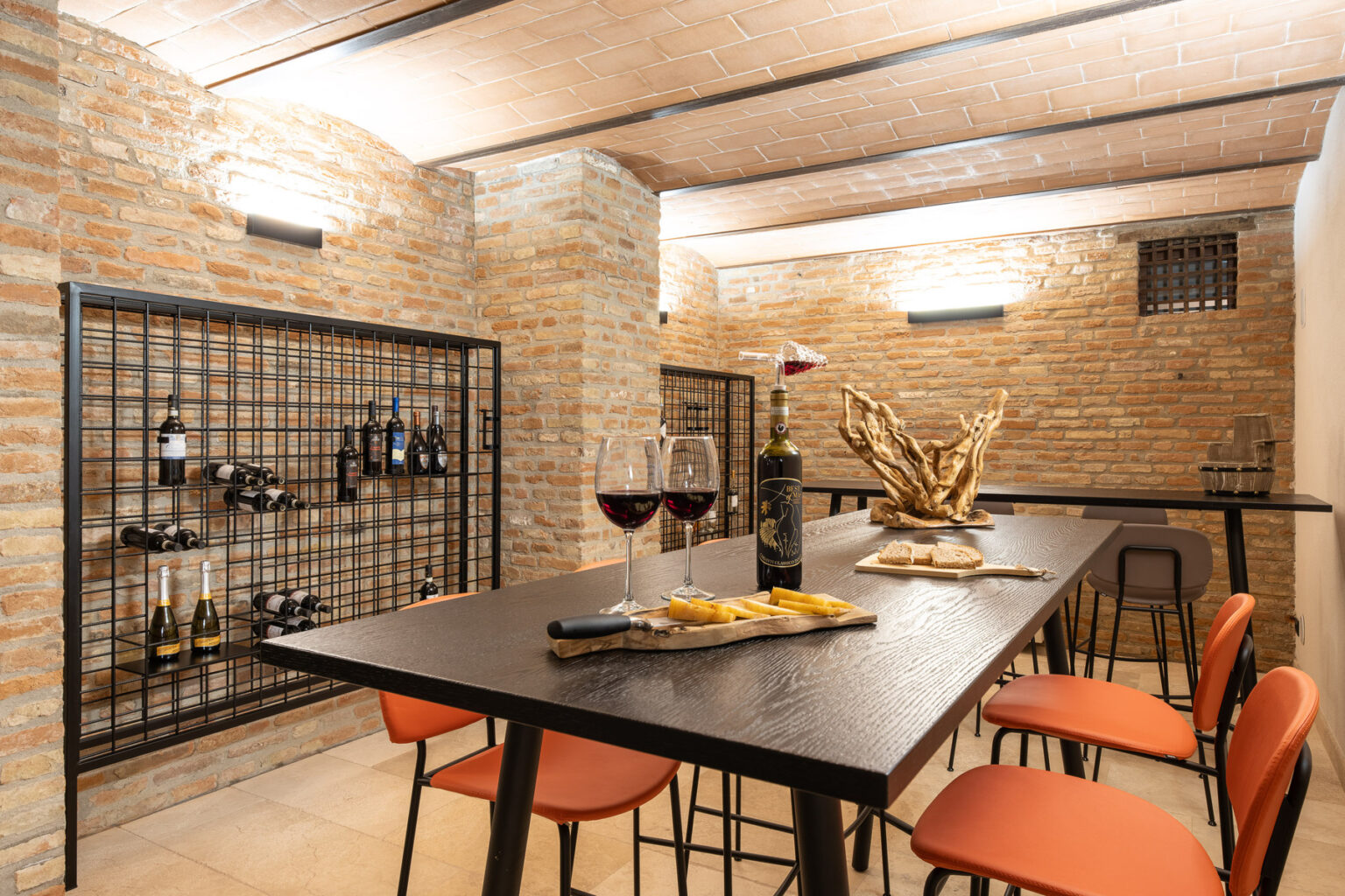 Modern wine cellar featuring a sleek black table, orange chairs, and a brick wall. The room includes wine storage racks and a decorative centerpiece, creating an inviting atmosphere for wine tasting and gatherings. | Only on SopranoVillas