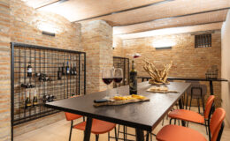 Modern wine cellar featuring a sleek black table, orange chairs, and a brick wall. The room includes wine storage racks and a decorative centerpiece, creating an inviting atmosphere for wine tasting and gatherings.