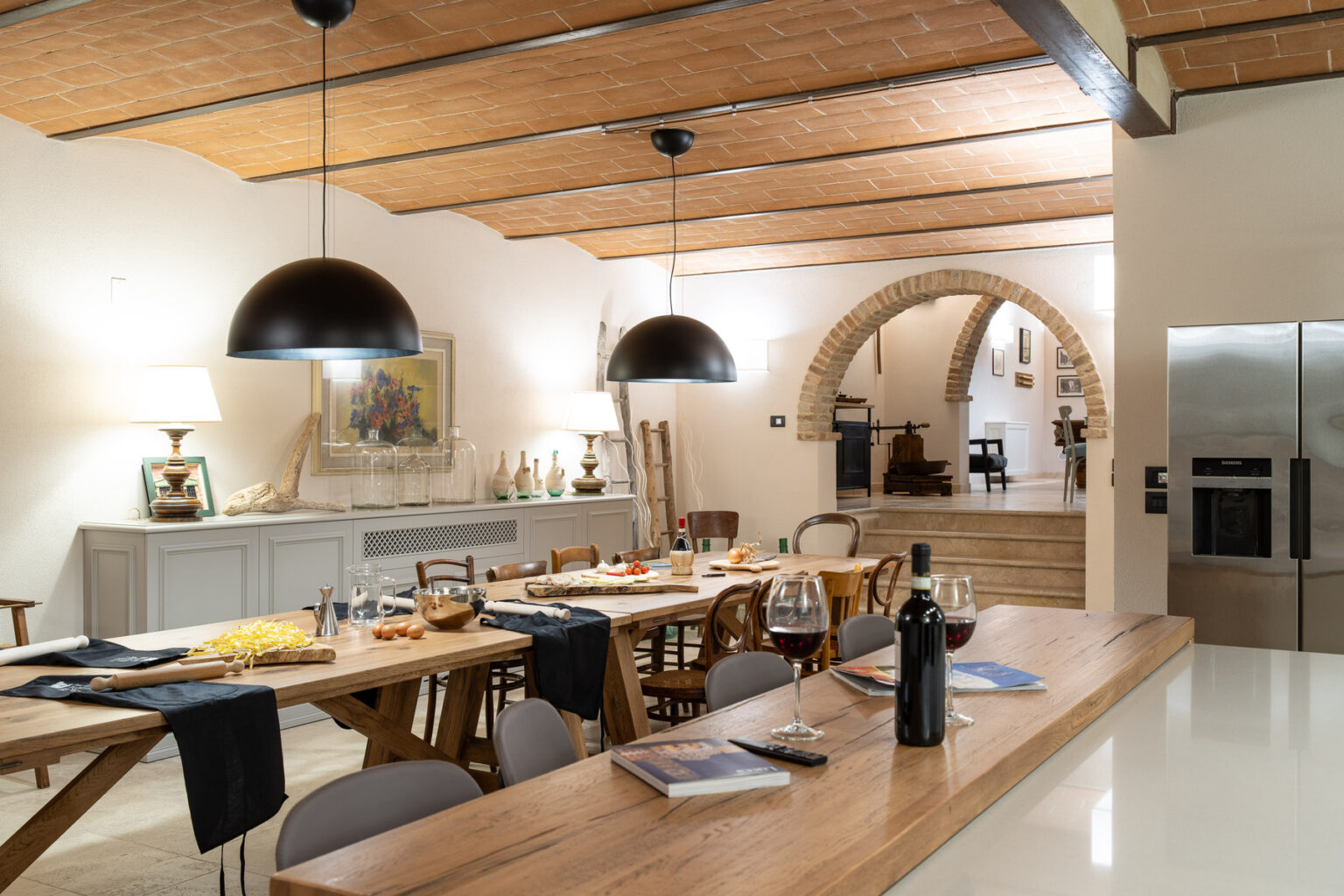 Modern dining area featuring a rustic wooden table set for a meal, with stylish black pendant lights and an open archway leading to a kitchen. The room has a warm ambiance, highlighted by natural wood beams and contemporary decor. | Only on SopranoVillas