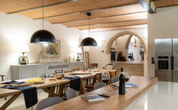 Modern dining area featuring a rustic wooden table set for a meal, with stylish black pendant lights and an open archway leading to a kitchen. The room has a warm ambiance, highlighted by natural wood beams and contemporary decor.