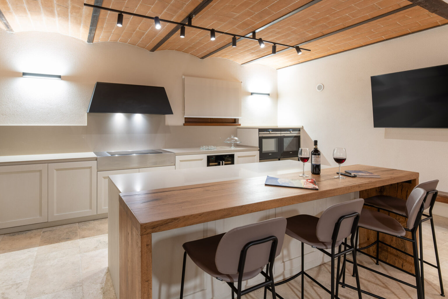 Modern kitchen interior featuring a spacious wooden island with bar stools, contemporary appliances, and warm lighting. A bottle of wine and glasses are set on the counter, enhancing the inviting atmosphere. | Only on SopranoVillas