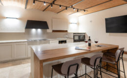 Modern kitchen interior featuring a spacious wooden island with bar stools, contemporary appliances, and warm lighting. A bottle of wine and glasses are set on the counter, enhancing the inviting atmosphere.
