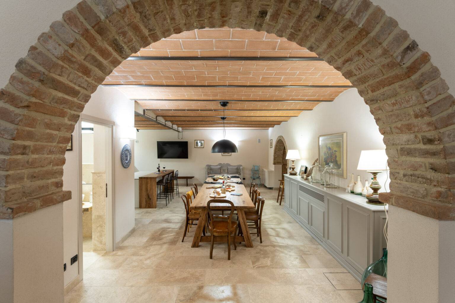 Spacious dining area with rustic wooden beams and an arched brick entrance, featuring a long wooden table surrounded by chairs, warm lighting, and tasteful decor elements in a cozy home setting. | Only on SopranoVillas