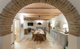 Spacious dining area with rustic wooden beams and an arched brick entrance, featuring a long wooden table surrounded by chairs, warm lighting, and tasteful decor elements in a cozy home setting.