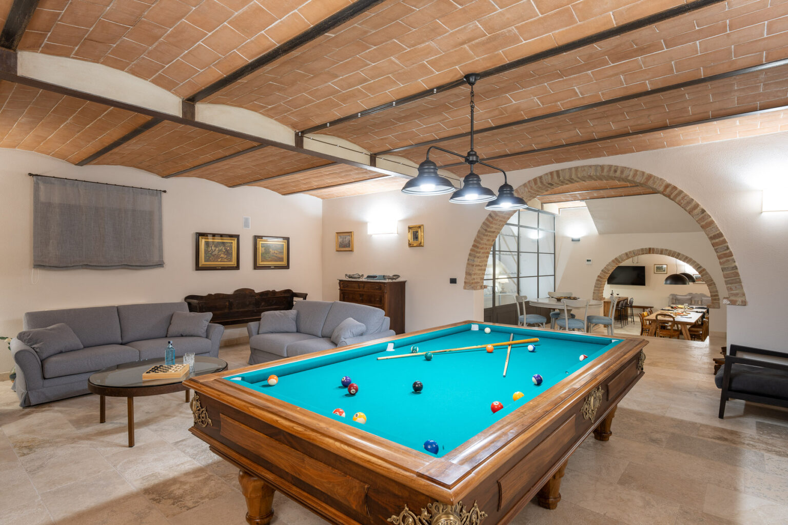 Spacious game room featuring a classic billiard table with teal felt, comfortable seating, and elegant decor, illuminated by stylish pendant lights and showcasing arched architectural details. | Only on SopranoVillas