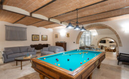 Spacious game room featuring a classic billiard table with teal felt, comfortable seating, and elegant decor, illuminated by stylish pendant lights and showcasing arched architectural details.