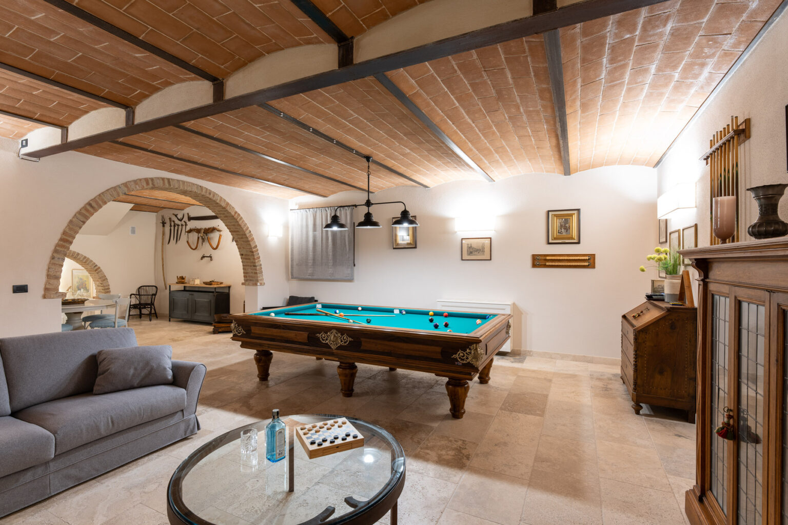 Spacious game room featuring a classic billiards table, cozy gray sofa, elegant wooden furniture, and stylish decor under a wooden beam ceiling, perfect for entertainment and relaxation. | Only on SopranoVillas