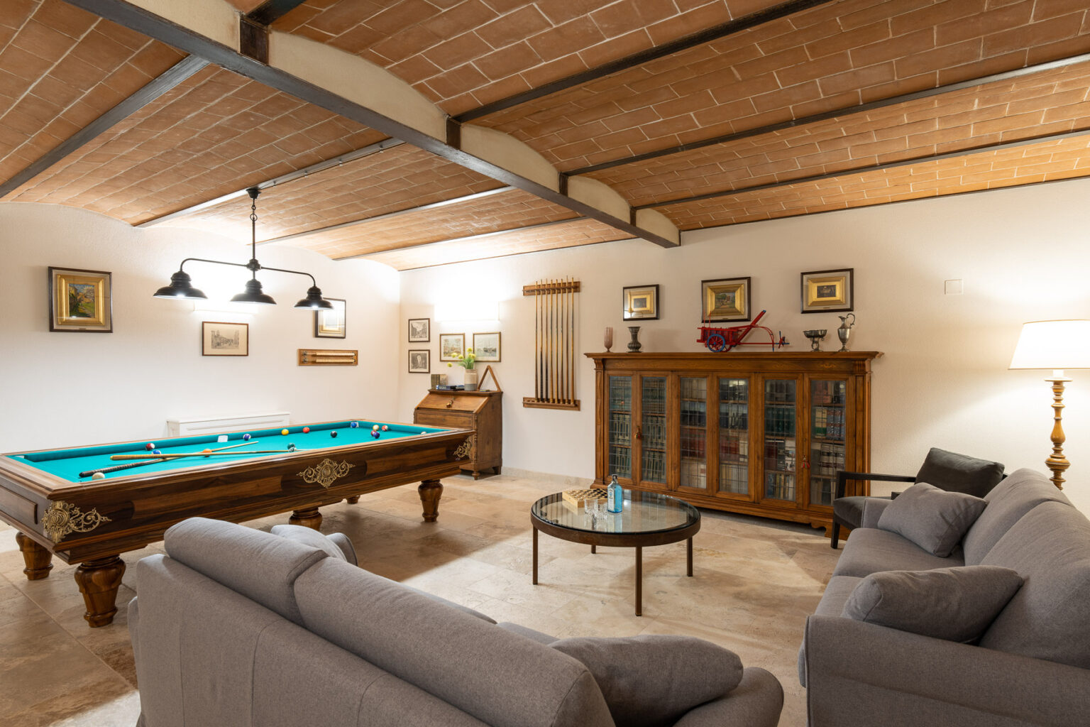 Cozy game room featuring a pool table, comfortable gray sofas, a glass coffee table, and wooden cabinetry, all under a stylish beamed ceiling. | Only on SopranoVillas