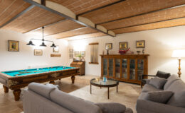 Cozy game room featuring a pool table, comfortable gray sofas, a glass coffee table, and wooden cabinetry, all under a stylish beamed ceiling.