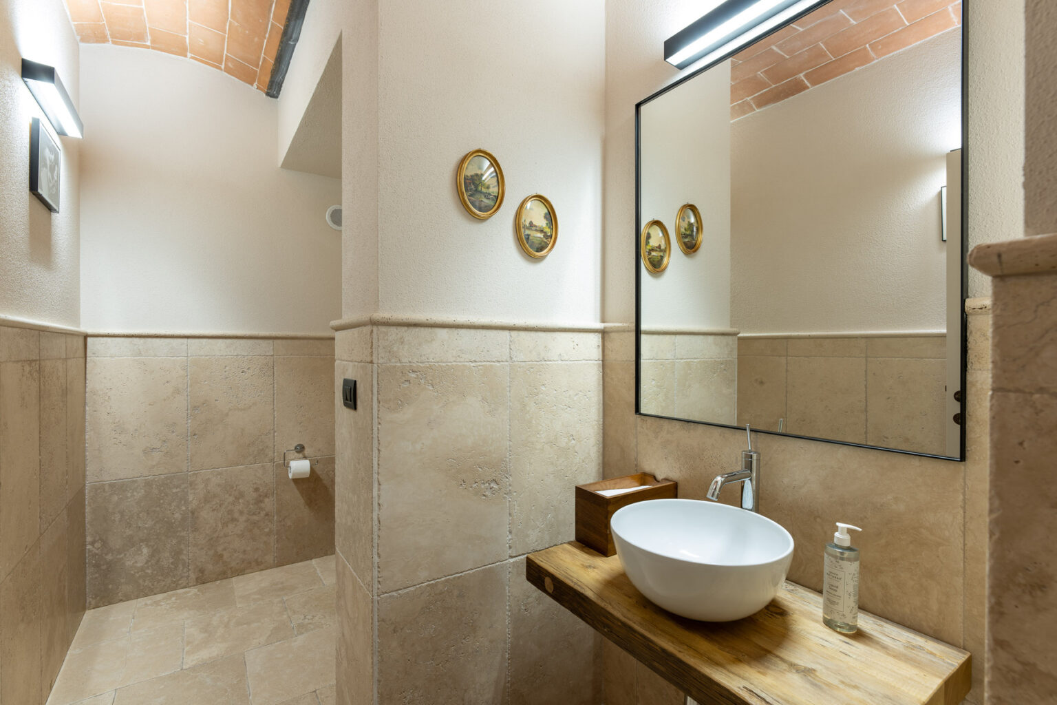 Modern bathroom interior featuring a curved ceiling, natural stone tiles, a stylish wooden countertop with a round basin, and decorative wall art. The space is illuminated by sleek lighting fixtures and includes essential amenities like a soap dispenser and toilet paper holder. | Only on SopranoVillas