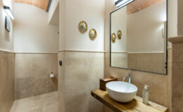 Modern bathroom interior featuring a curved ceiling, natural stone tiles, a stylish wooden countertop with a round basin, and decorative wall art. The space is illuminated by sleek lighting fixtures and includes essential amenities like a soap dispenser and toilet paper holder.