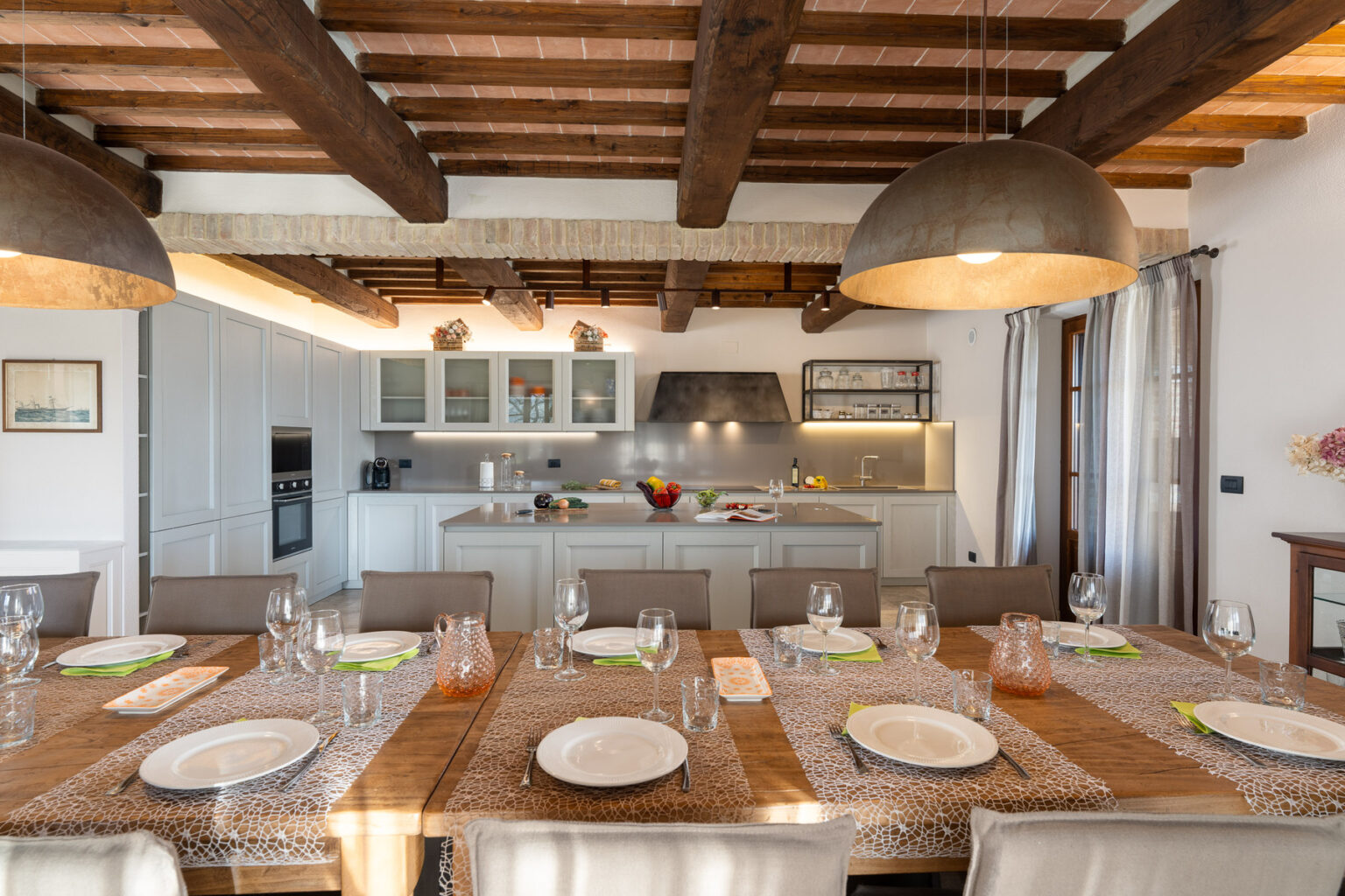 Modern dining room with a long wooden table set for a meal, featuring elegant tableware and glasses. The kitchen in the background showcases contemporary cabinetry and appliances, illuminated by stylish pendant lights and natural light from large windows. | Only on SopranoVillas