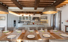 Modern dining room with a long wooden table set for a meal, featuring elegant tableware and glasses. The kitchen in the background showcases contemporary cabinetry and appliances, illuminated by stylish pendant lights and natural light from large windows.