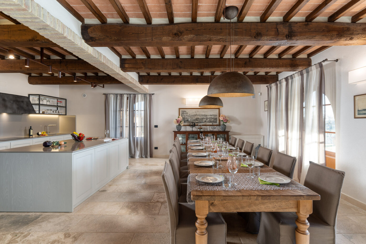 Modern dining room featuring a long wooden table set for a meal, surrounded by upholstered chairs. The space includes a sleek kitchen island with fresh fruits, pendant lighting, and large windows adorned with sheer curtains, highlighting a warm and inviting atmosphere. | Only on SopranoVillas
