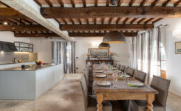 Modern dining room featuring a long wooden table set for a meal, surrounded by upholstered chairs. The space includes a sleek kitchen island with fresh fruits, pendant lighting, and large windows adorned with sheer curtains, highlighting a warm and inviting atmosphere.