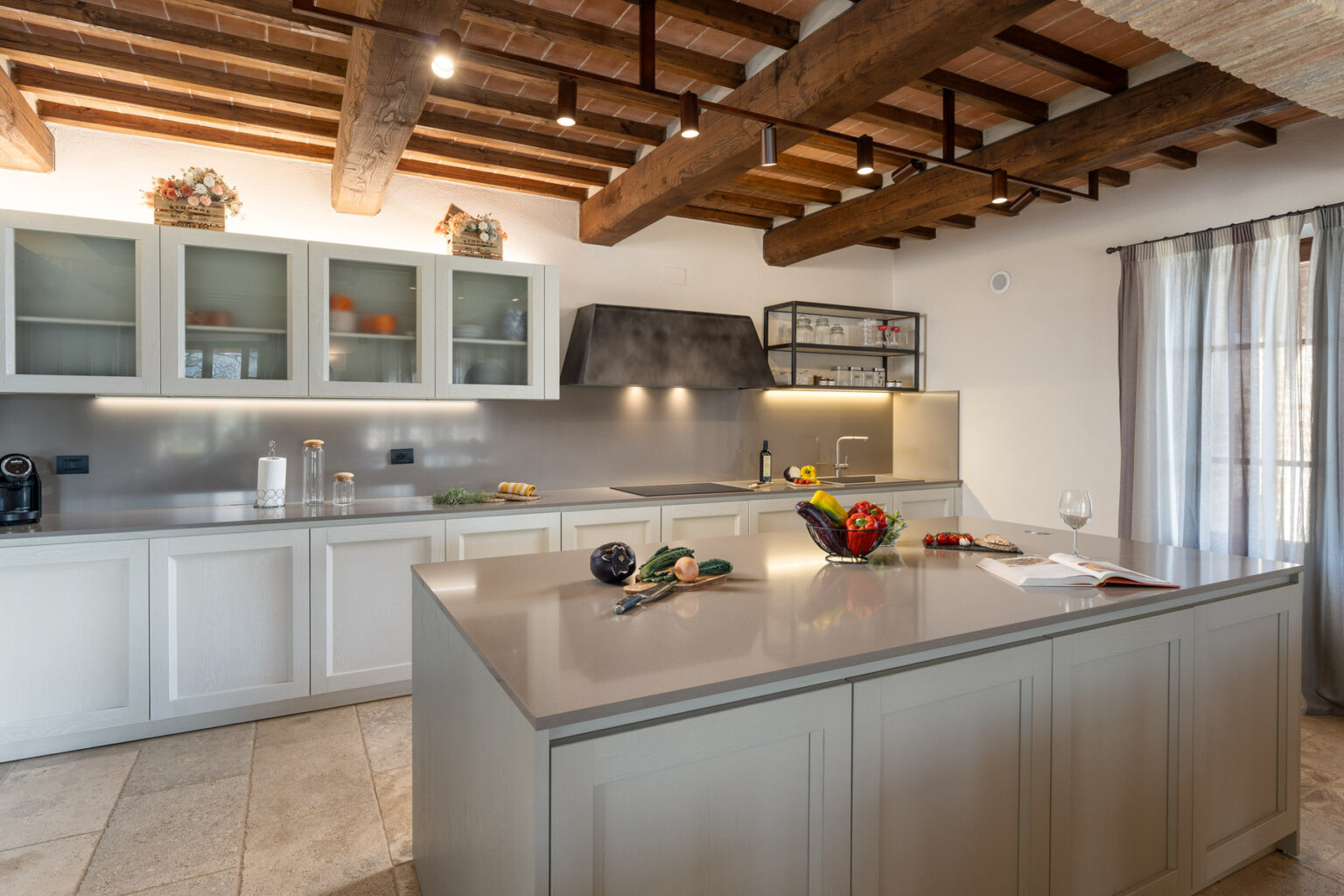 Modern kitchen interior featuring a spacious island with fresh vegetables, a stylish countertop, wooden beams, and elegant cabinetry. Natural light streams through the window, enhancing the inviting atmosphere. | Only on SopranoVillas